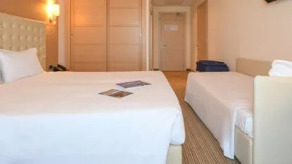 Camera d'albergo con due letti singoli, biancheria bianca, armadio in legno e valigia blu sul pavimento.