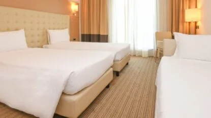 Camera d'hotel luminosa con tre letti singoli, materassi bianchi, testiera imbottita e tende beige.