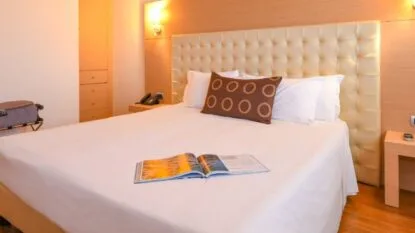 Camera d'albergo con letto matrimoniale, testata imbottita beige, cuscino marrone, rivista aperta e luci calde ai lati.