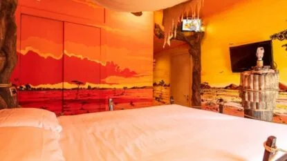 Camera decorata con murales arancioni e rossi di paesaggi africani, letto bianco e TV a parete.