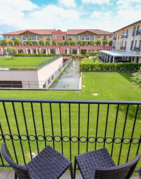Balcone con sedie di vimini e vista su giardino verde e edificio residenziale con panchine e fontane.