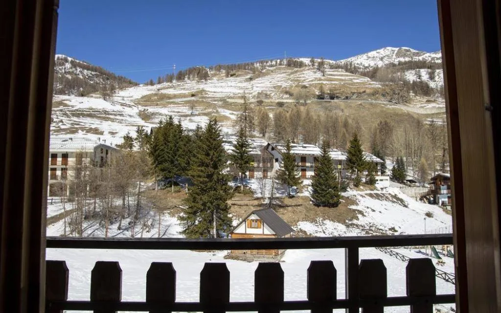 Vista dal balcone su montagne innevate, pini e chalet alpini sotto cielo azzurro