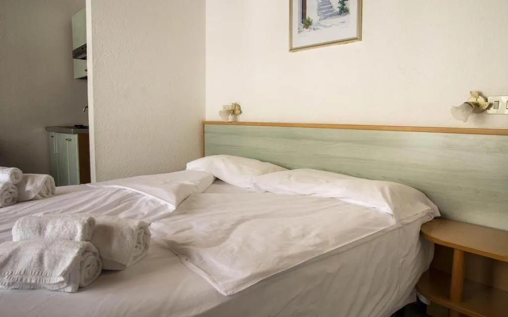 Letto matrimoniale con testiera verde, asciugamani arrotolati e angolo cottura sullo sfondo