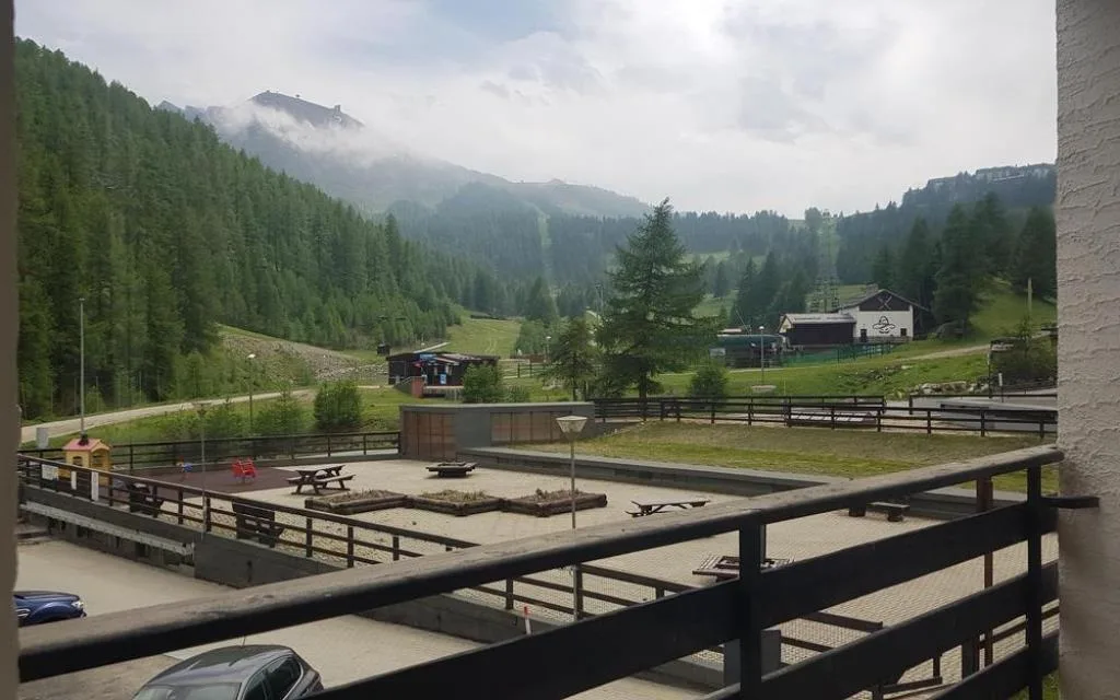 Vista dal balcone su piazzale, area giochi e montagne verdi con nebbia