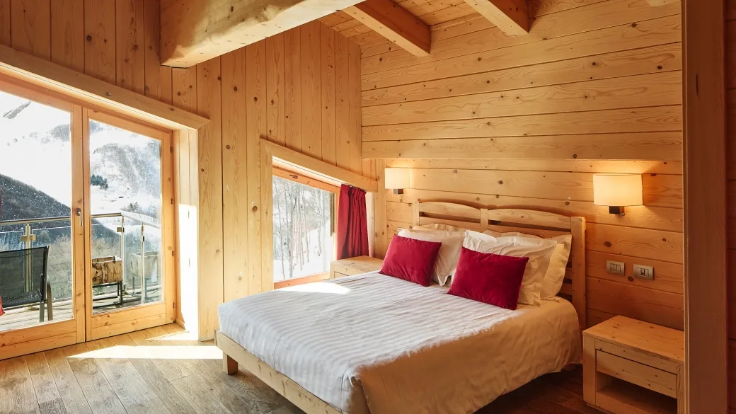 Camera matrimoniale in legno con cuscini rossi e ampia vetrata sulle montagne