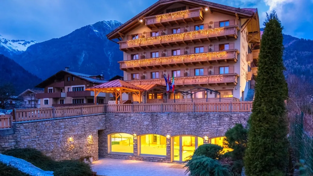 Facciata dell'hotel al crepuscolo con luci calde e area benessere illuminata al piano terra