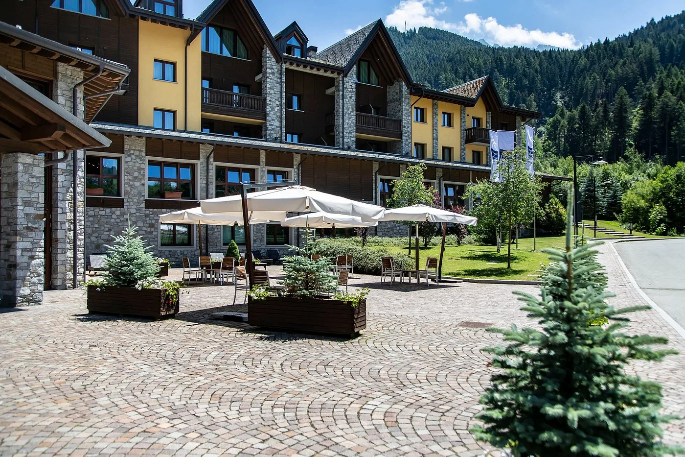 Cortile dell'hotel in estate con ombrellone, tavoli, abeti in vaso e pavimentazione in pietra