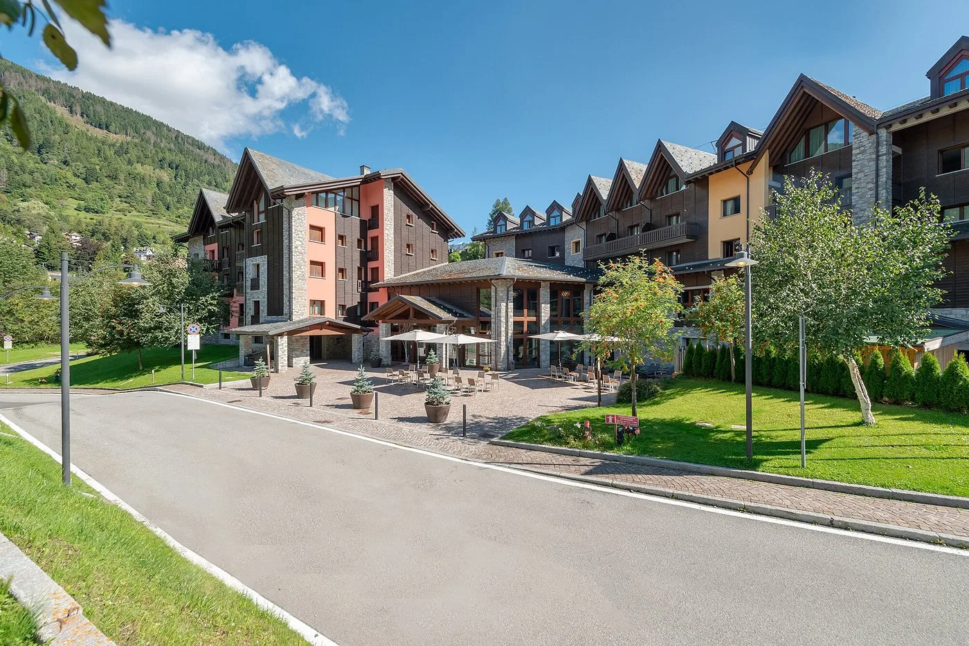 Esterno estivo dell'hotel con ingresso, ombrelloni e giardini verdi ai piedi delle montagne
