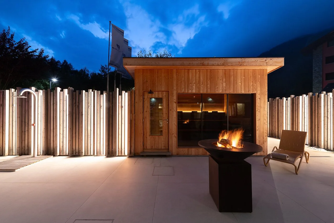 Sauna esterna in legno illuminata al crepuscolo con braciere acceso e doccia emozionale
