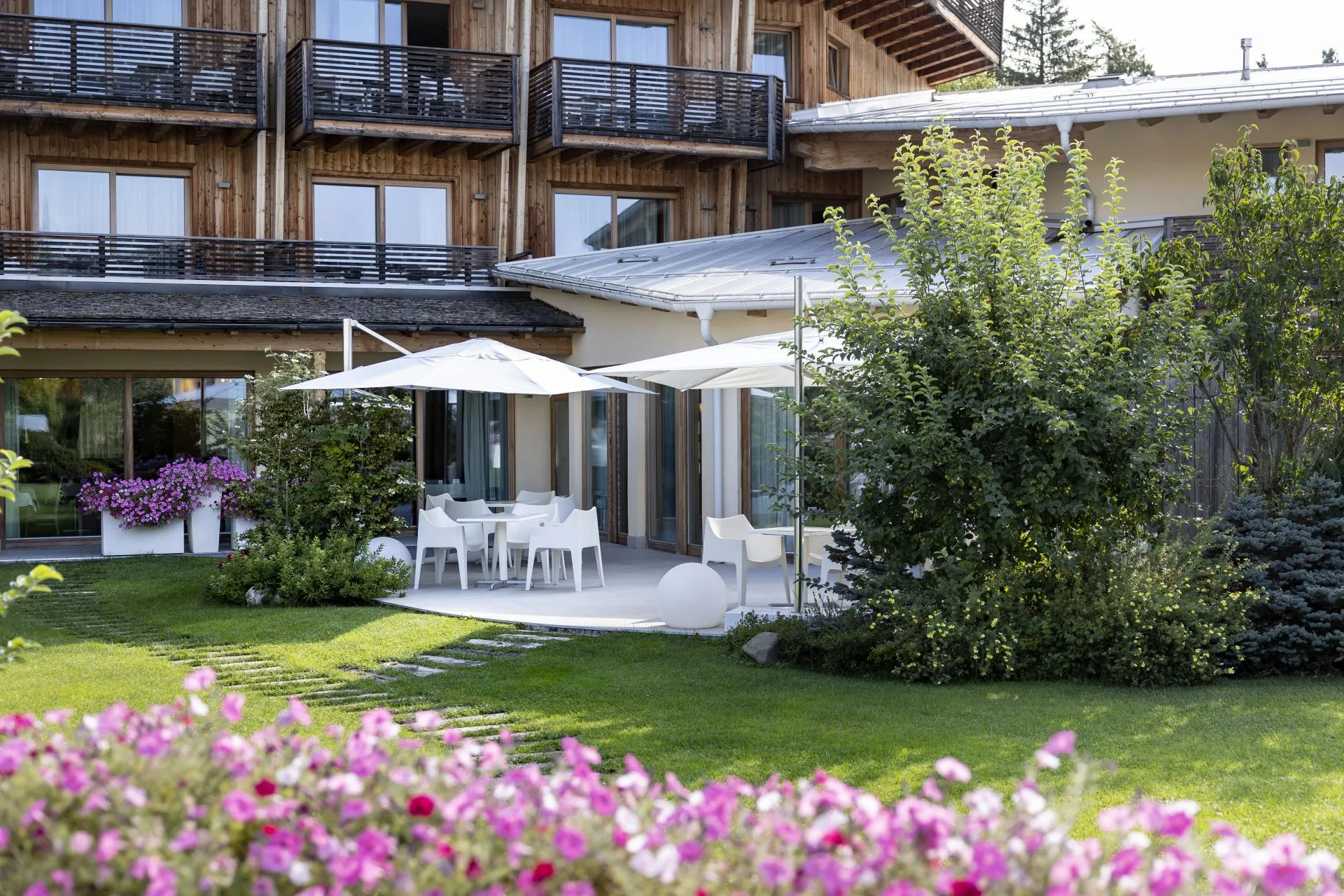 Giardino estivo fiorito con terrazza, tavoli bianchi, ombrelloni e facciata in legno