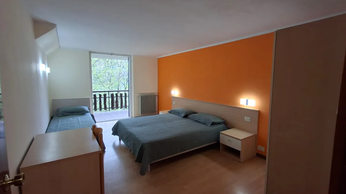 Camera tripla con letto matrimoniale e singolo, parete arancione e accesso al balcone con vista sul bosco