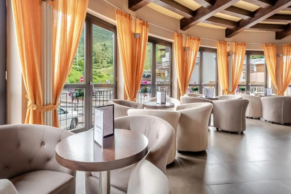 Sala lounge bar con poltrone bianche, tavolini rotondi, tende arancioni e vista sulle montagne
